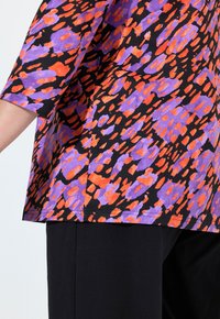 Lyhythihainen paita, jossa on musta kangas ja värikäs abstrakti kuvio oranssina ja purppurana. Soljuva muotoilu, rento istuvuus ja yksinkertainen helma.