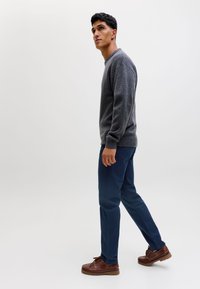 Mand går iført en grå sweater, marineblå bukser og brune læder sko mod en ensartet lys baggrund.