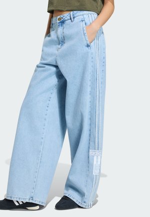 Person trägt hellblaue Jeans mit weitem Bein, Adidas-Logo und Streifen an der Seite, kombiniert mit schwarzen Adidas-Sneakern.