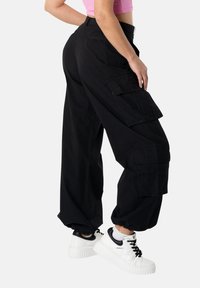 Henkilö, jolla on mustat taktiset housut ja valkoiset korolliset lenkkarit, seisoo yhdellä jalalla hieman koukistettuna, yksiväristä taustaa vasten.