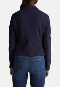 Kvinna som bär en marinblå kavaj och blå denimjeans, visad bakifrån mot en vit bakgrund.