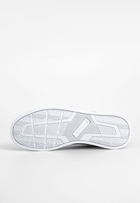 Semelle blanche de basket avec motif de bande de roulement texturé et une section rectangulaire avec logo au centre, sur un fond blanc uni.