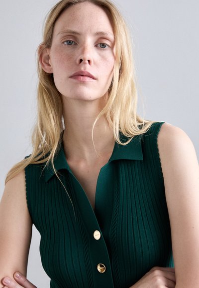 Blonde vrouw met neutrale uitdrukking draagt een mouwloos groen ribtop met gouden knopen, staande tegen een effen lichtgrijze achtergrond.