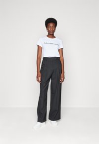 Ontspannen-grijze broek met hoge taille en plooien, gecombineerd met een wit katoenen t-shirt met de tekst "Calvin Klein Jeans". Witte sneakers.