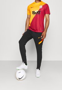 Strój piłkarski z czerwoną i pomarańczową koszulką z logo sponsora, w połączeniu z czarnymi spodniami i białymi korkami, stojący obok białej piłki.