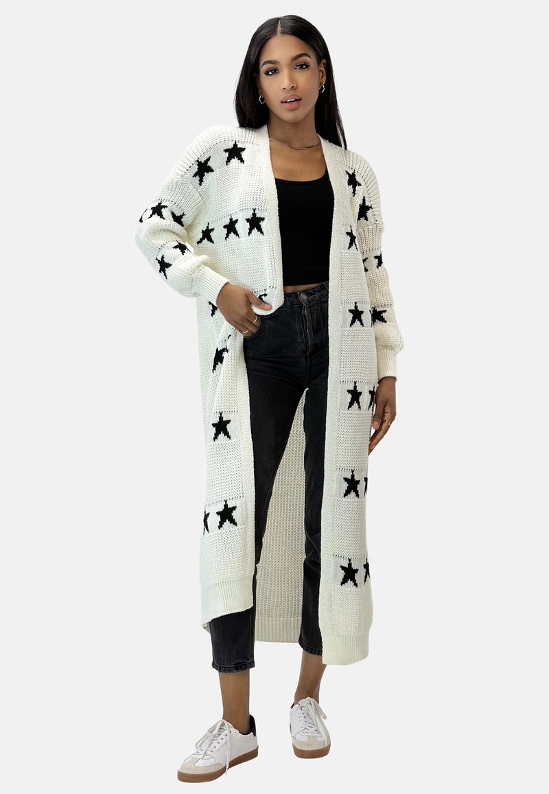 Cardigan lungo bianco lavorato a maglia con motivi a stella nera, apertura frontale, polsini a coste e tasche laterali, indossato sopra un top nero e jeans.