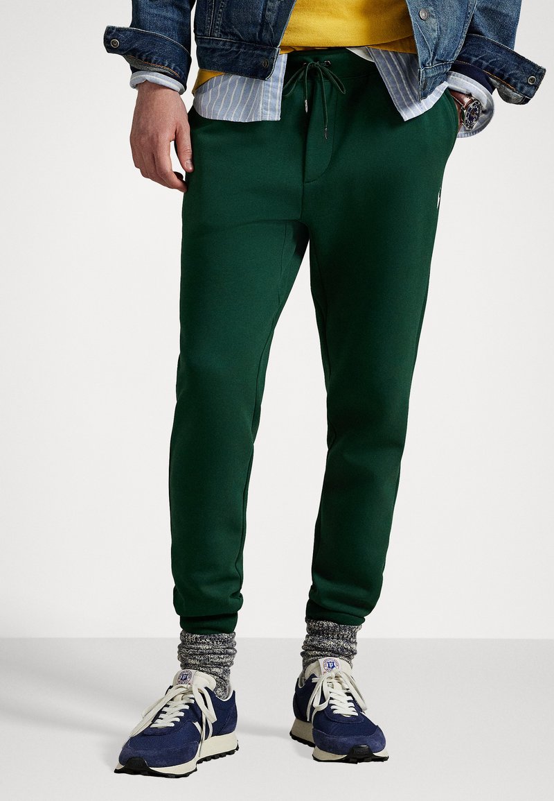 Grønne joggebukser i et elastisk stoff, med snøring i midjen, sidelommer og tettsittende mansjetter. Kombinert med joggesko og mønstrede sokker.