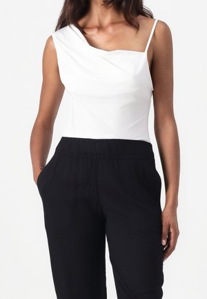 Mujer con una blusa blanca asimétrica sin mangas, con una tirante delgada, y pantalones negros de cintura alta con bolsillos.