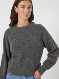Mulher vestindo um suéter macio, felpudo e cinza escuro, com mangas compridas e jeans azuis, em pé com uma mão no bolso contra um fundo liso.