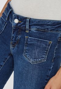 Högmidjade blå denimjeans med femfickorsdesign, metallknappstängning och orange sömmar på en slät, texturerad tyg.