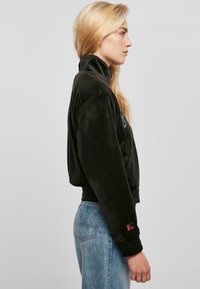 Chaqueta corta de terciopelo negro con cuello alto, puños acanalados y cinturilla. Presenta un discreto detalle del logo en el pecho. Combinada con jeans azules.