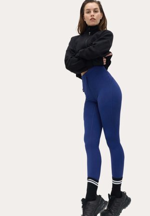 Ung kvinde stående med armene krydsede iført en sort cropped jakke, blå højtaljede leggings, stribede strømper og sorte sneakers.
