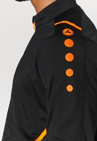 Svart idrottsjacka med hög krage, har orange detaljer, inklusive en rand och cirkulära inslag längs ärmen. Slät textur.