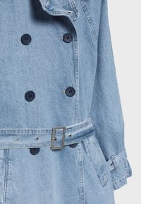 Trench-coat en denim bleu clair avec boutons noirs doubles, ceinture réglable avec boucle métallique et sangles aux poignets.
