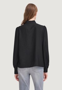 Blusa nera con scollo alto, maniche lunghe a sbuffo e vestibilità ampia. Realizzata in un tessuto liscio, indossata con jeans grigi chiari.