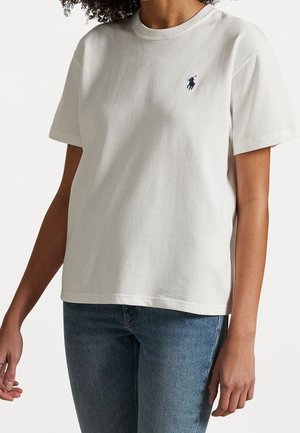 Woman wearing a plain white short-sleeve T-shirt with a small dark logo on the left chest and blue jeans.