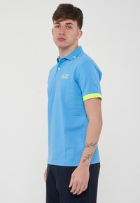 Polo shirt azzurro chiaro a maniche corte, con dettagli verde neon sul colletto e sulle maniche, e un logo sul lato sinistro del petto.