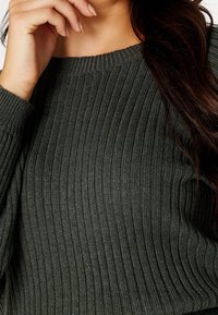 Femme aux longs cheveux bruns portant un pull côtelé vert foncé, main près du visage, cadrée au niveau du cou et de la taille.