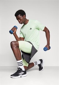 Grünes Sport-T-Shirt mit schwarzem Logo, passende mintfarbene Shorts, schwarze Schuhe mit weißen Akzenten, hält eine blaue Hantel, kniet auf einem Bein.