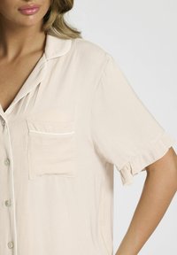 Korte mouw beige button-up shirt van zachte stof met een enkele borstzak, witte bies langs de kraag en mouwranden, en een gerimpelde zoom.