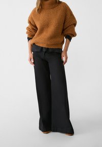 Femme portant un pull en maille épaisse marron, un pantalon large noir et des chaussures marron, debout devant un fond clair uni.