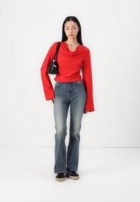 Rød langermet topp med båtutringning, sammen med blå denimjeans med vide ben, og svarte joggesko. Modellen holder en liten svart håndveske.
