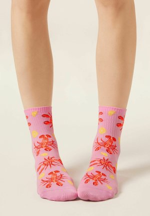 Nogi w różowych skarpetkach z czerwonymi wzorami homarów i żółtymi plasterkami cytryny, stojące na jasnym neutralnym tle.