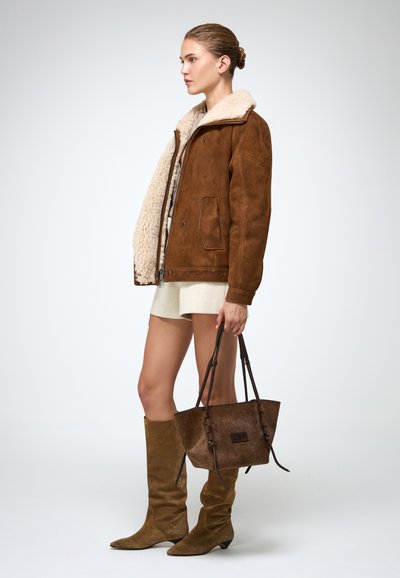 Bruine schapenvachtjas met een crèmekleurige kraag, gecombineerd met beige shorts, lange bruine suède laarzen en een bruine tote bag.