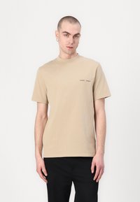 Beige színű pamut póló, rövid ujjakkal és kerek nyakkivágással. A mellkason kis nyomtatott szöveg található. Fekete nadrággal viselve.