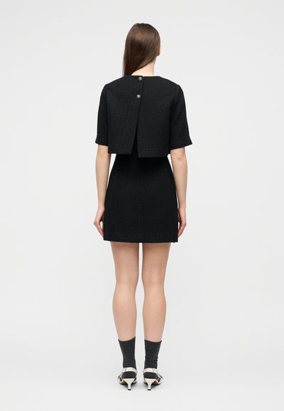 Γυναίκα που στέκεται πλάτη, φορώντας μαύρο φόρεμα με κοντά μανίκια και κοντή επικάλυψη, καθώς και λευκά παπούτσια με ψηλό τακούνι και σκούρες κάλτσες.