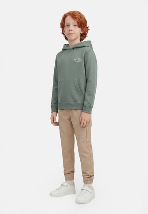 Jongen met rood haar staand, draagt een groen sweatshirt met capuchon, beige cargobroek en witte sneakers op een effen witte achtergrond.