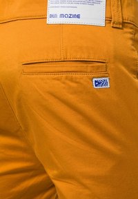 Oranje katoenen broek met een achterzak voorzien van een blauw logolabel en een klein patchlabel erboven, gestikt met contrasterend garen.