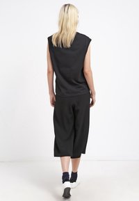 Blonde vrouw die wegloopt in een mouwloos zwart topje, brede zwarte cropped broek, marineblauwe sokken en witte sneakers, op een witte achtergrond.