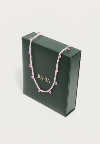 Collier délicat avec de petites perles blanches rondes et des perles en forme de goutte rose, posé sur une boîte vert foncé avec des lettres dorées.