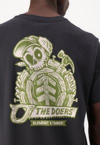 Black cotton t-shirt featuring a green graphic design of a skull with tools, surrounded by wood patterns, and the text "THE DOERS ELEMENT X TIMBER!"
