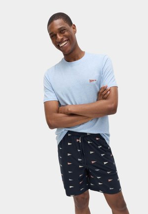 Uomo sorridente in piedi con le braccia incrociate, indossa una maglietta azzurra e pantaloncini blu navy decorati con piccoli bandierine colorate.