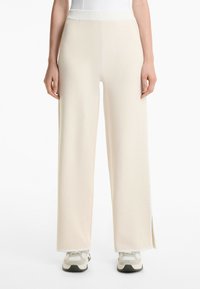 Pantalons à jambes larges en tissu crème, présentant une taille haute, une texture côtelée et des fentes sur les côtés. Assortis à des baskets gris clair et blanches.