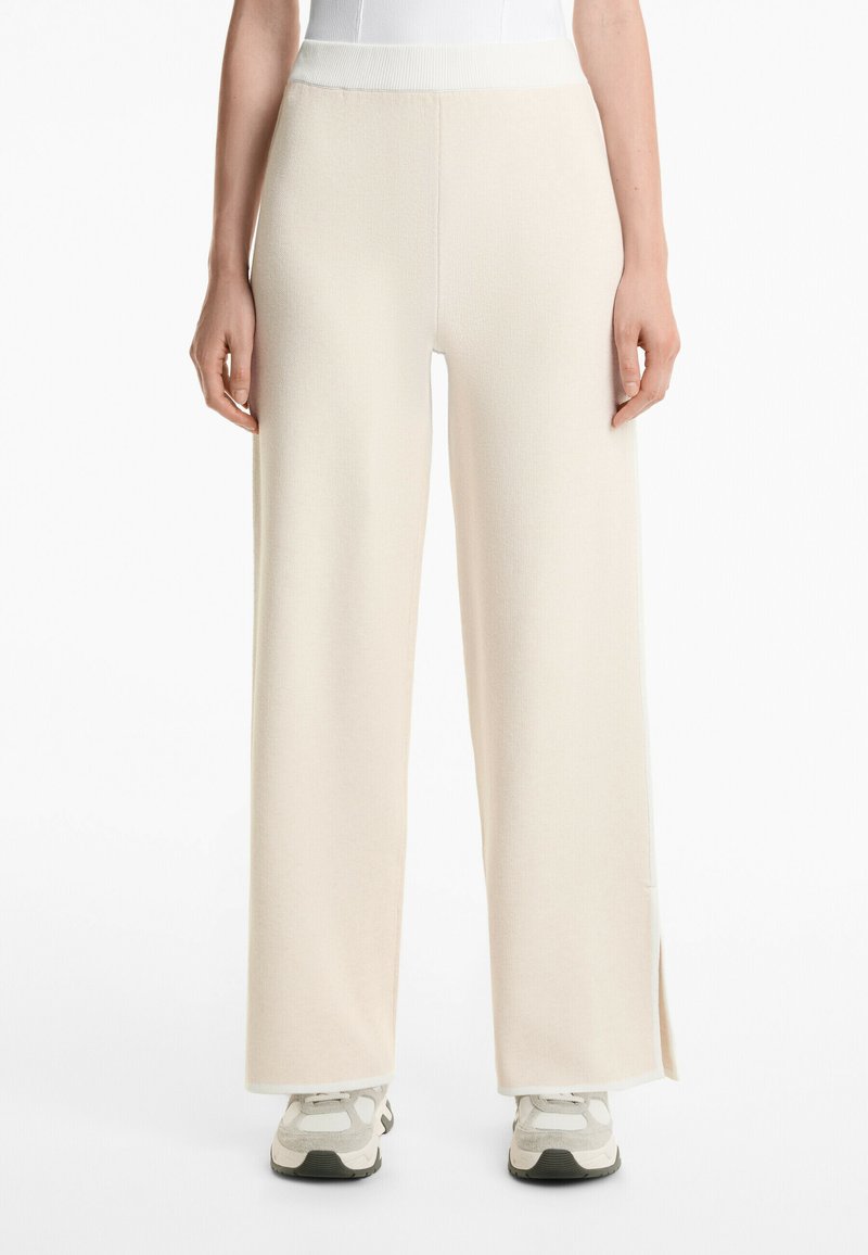 Pantalons à jambes larges en tissu crème, présentant une taille haute, une texture côtelée et des fentes sur les côtés. Assortis à des baskets gris clair et blanches.