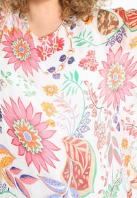 Persona con cabello rizado que lleva una blusa blanca con grandes patrones florales en colores rosa, naranja, verde, púrpura y azul.