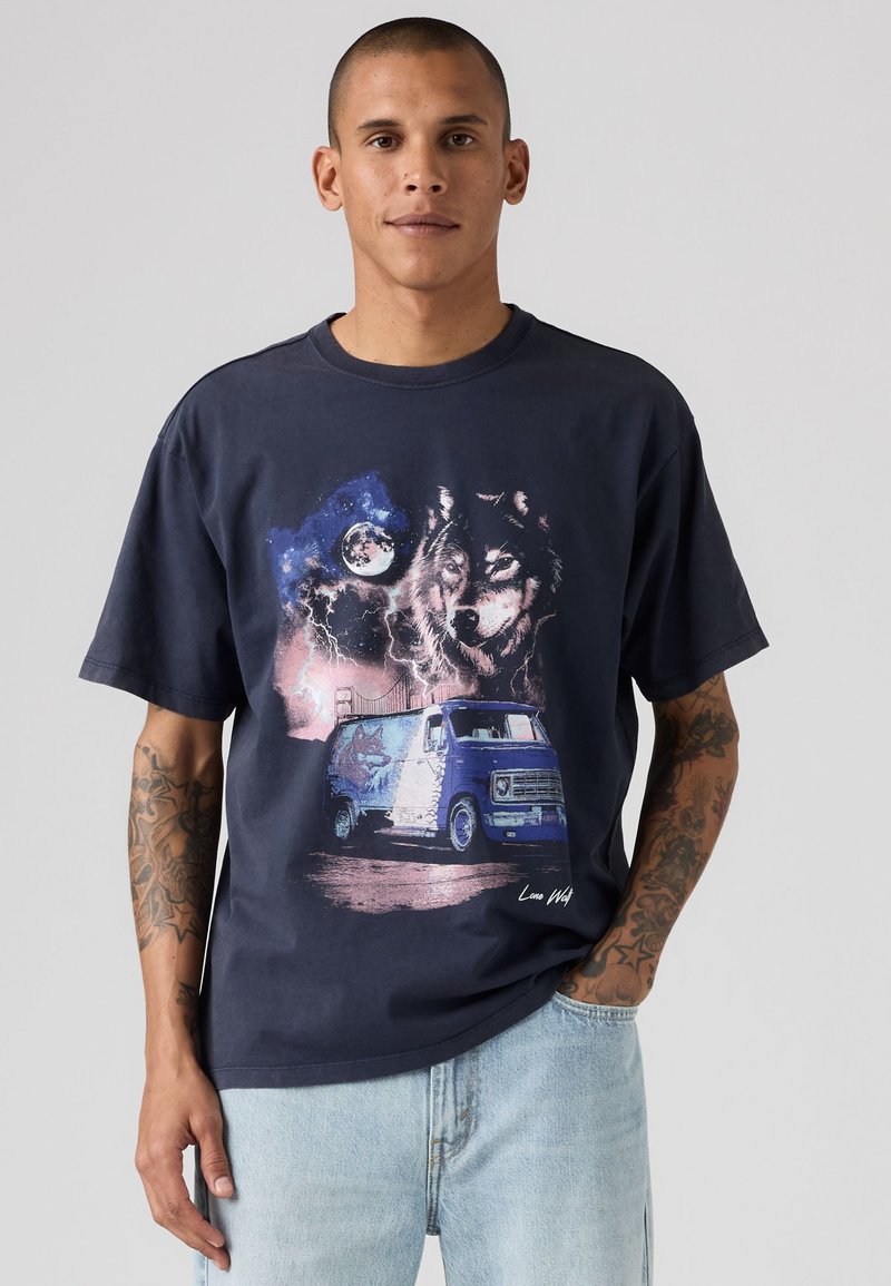 Uomo che indossa una t-shirt blu navy con grafica di lupo, luna piena, fulmine, ponte e furgone blu, abbinata a jeans azzurri chiari, in piedi su sfondo grigio.