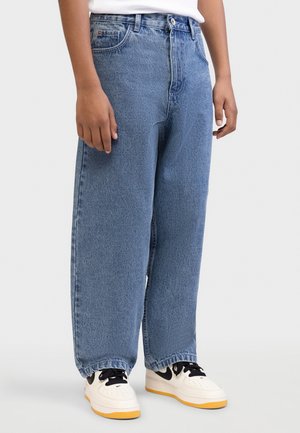 Persoană purtând blugi largi albaștri și adidași albi cu accente negre, stând în picioare pe un fundal simplu, deschis la culoare.