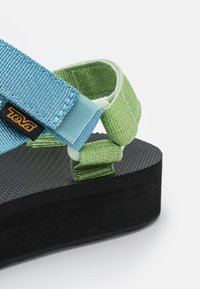 Teva-sandaalit ovat varustettu mustalla kumipohjalla, teallanssin ja vihreän värisillä kudotuilla hihnoilla, säädettävillä soljilla ja tekstuurilla varustetulla pohjalla tuen tarjoamiseksi.