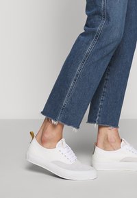 Zapatillas de lona blancas con un diseño texturizado, parte delantera con cordones y una pestaña de acento amarilla en el talón, combinadas con jeans de denim azul cortos.