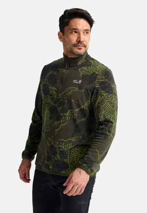 Mann trägt eine grün-schwarze gemusterte Jack Wolfskin Fleecejacke und schwarze Hose, schaut seitlich mit neutralem Gesichtsausdruck.