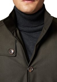 Ciemnooliwkowa kurtka z wysokim kołnierzem, zestawiona z ciemnoszarym golfem. Kurtka ma detal w postaci guzików na kołnierzu.