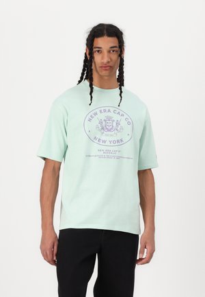Tricou din bumbac verde menta cu mâneci scurte, având un imprimeu grafic purpuriu cu "New Era Cap Co." și detalii într-un design circular.