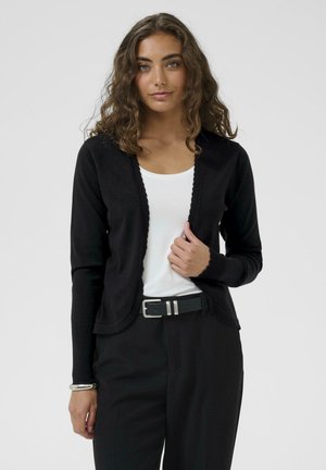 Vrouw met krullend bruin haar die een zwarte cardigan draagt over een wit topje, zwarte broek en een zwarte riem, staand voor een effen achtergrond.