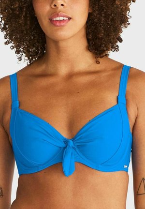 Vrouw met krullend haar die een felblauwe bikinitop draagt met een knoop in het midden en verstelbare bandjes tegen een effen achtergrond.