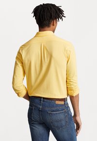 Hombre con rastas que lleva una camisa de manga larga amarilla y jeans azules, de pie con la espalda hacia la cámara frente a un fondo simple.