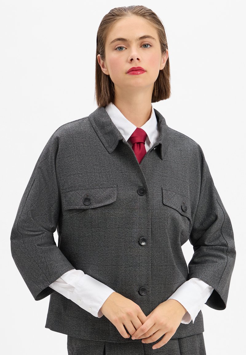 Graue karierte Überjacke mit kragenförmigem Ausschnitt, zwei Brusttaschen und schwarzen Knöpfen. Darunter sichtbar ist ein weißes Hemd und eine rote Krawatte.